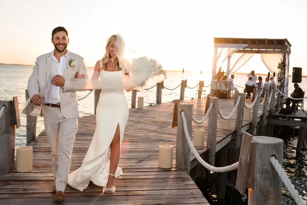 Bride and groom walking joyfully down a pier with guests and sunset in the background after their beachside ceremony, captured by a renowned wedding photographer in Cancun.