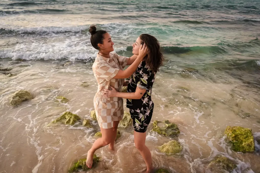 Two women share a playful moment by the seaside, with waves lapping at their feet and the glow of the horizon in the background, captured by a family photographer in Cancun.