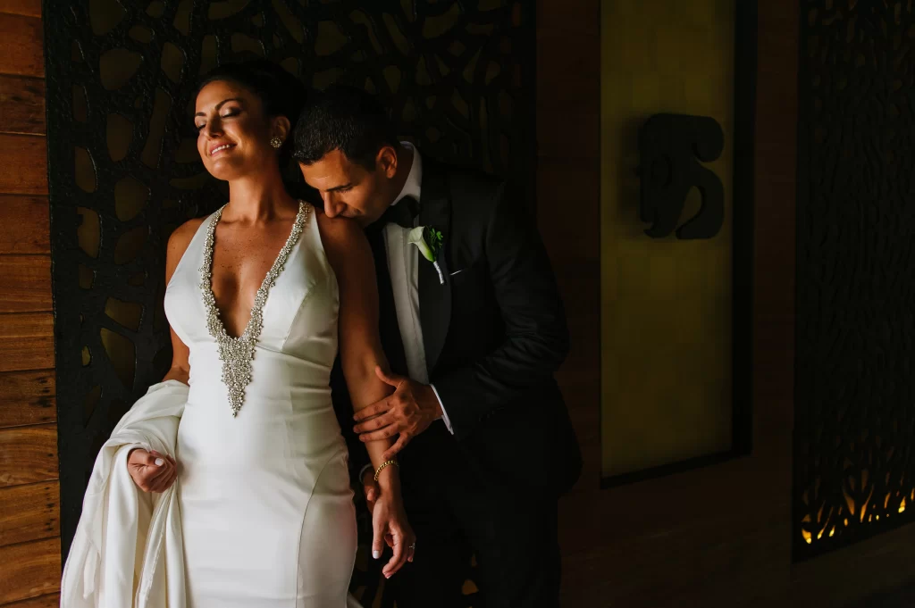 A tender moment as a groom lovingly embraces his bride from behind, both smiling and radiating happiness on their wedding day, against a warm, softly lit wooden backdrop. Captured by an experienced wedding